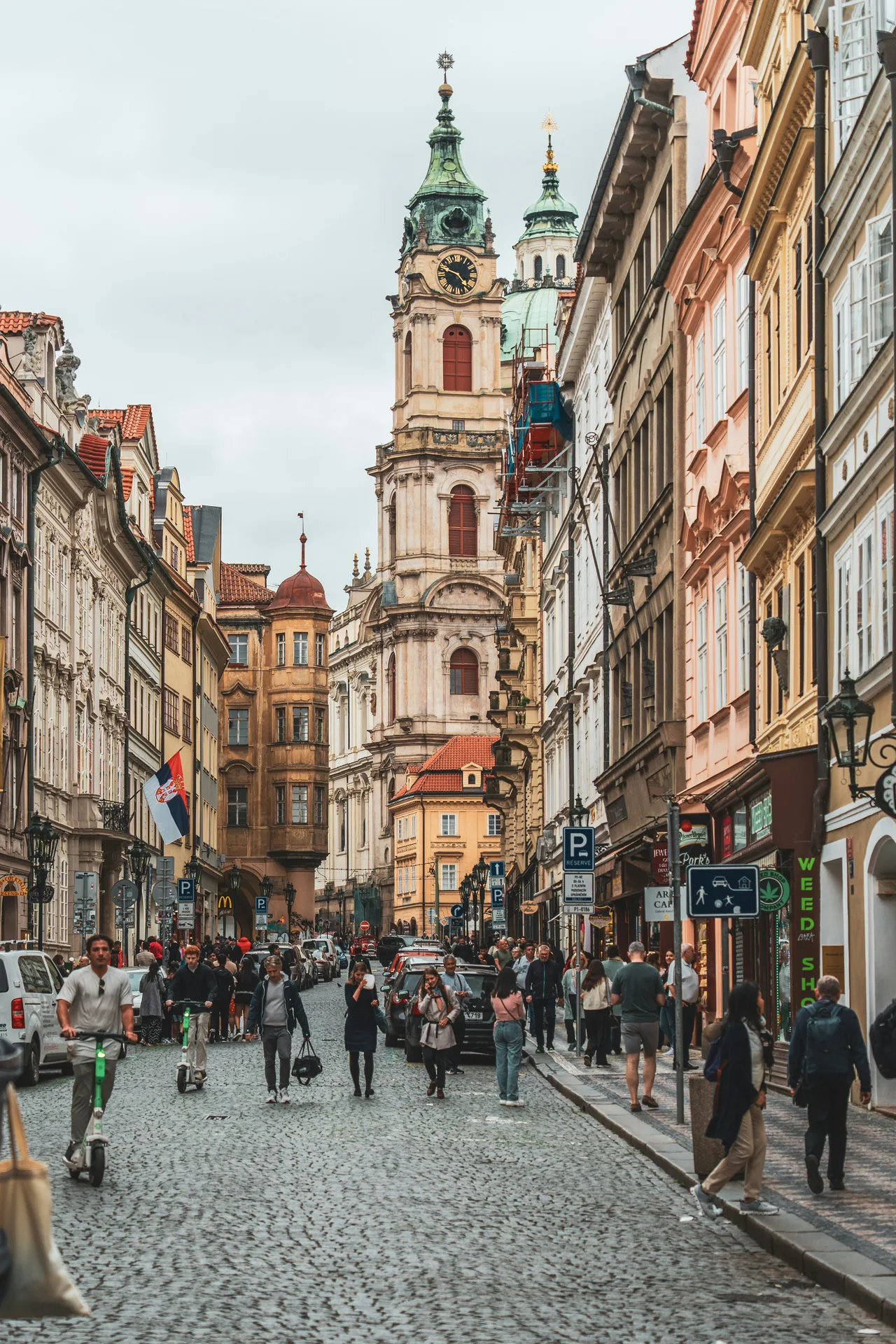 Dlážděná pražská ulice s historickými fasádami a kostelní věží.